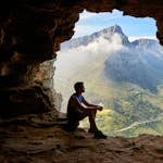 A man sitting in a cave overlooking a majestic mountain landscape under daylight.