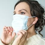 Adult woman adjusting blue surgical mask indoors for health safety.